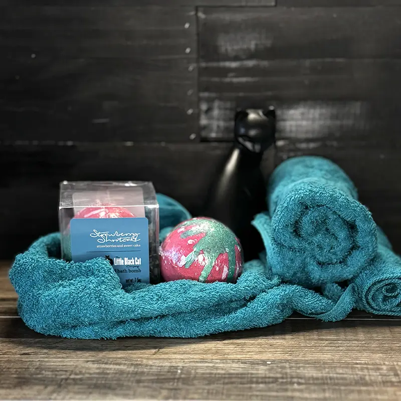 artistic arrangement of one boxed and one loose bath bomb with a towel and an art deco cat statue